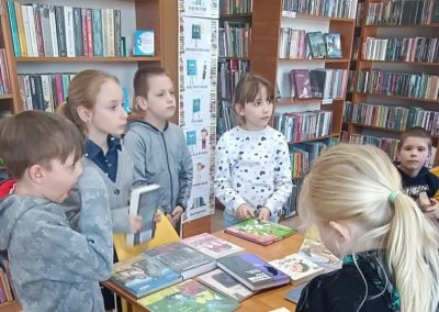 Grupa kilku dzieci stoi przy stole w bibliotece, oglądając ułożone na nim książki. W tle widać wysokie regały wypełnione książkami oraz oznaczenia działów. Atmosfera przypomina zajęcia edukacyjne lub wspólne wybieranie lektur.