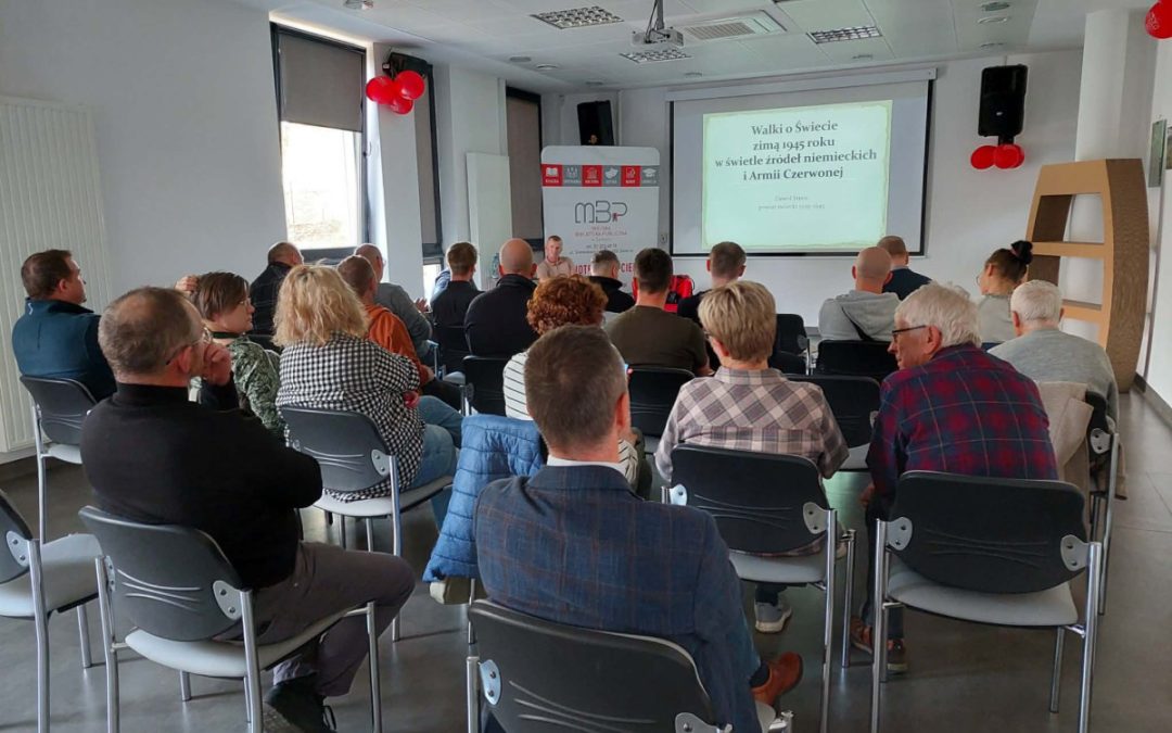 W szarej sali na krzesłach siedzą widzowie i prowadzący zajęcia. Na ścianie wyświetla się slajd z tekstem. W sali jest też tablica z logo MBP w Świeciu.