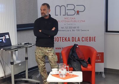 Prelegent stoi z założonymi rękami obok czerwonego fotela i stolika z wodą podczas spotkania autorskiego w bibliotece. W tle znajduje się baner Miejskiej Biblioteki Publicznej w Świeciu.