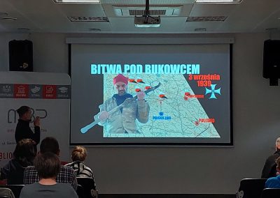 Na ekranie wyświetlony jest slajd zatytułowany „Bitwa pod Bukowcem 3 września 1939”. Obok ekranu stoi prowadzący prezentację, a uczestnicy siedzą w zaciemnionej sali bibliotecznej.