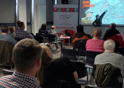 Prelegent wskazuje mapę na dużym ekranie podczas wykładu zatytułowanego „Armia Pomorze”. Publiczność siedzi w rzędach i obserwuje prezentację.
