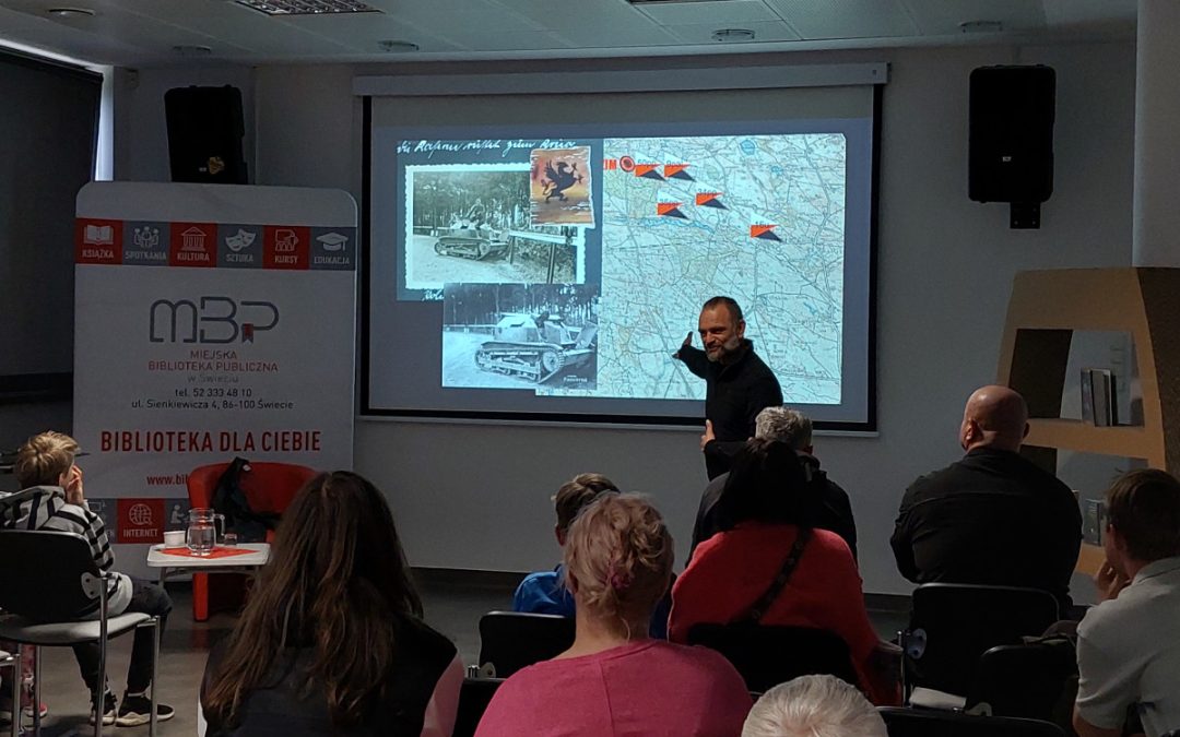 Prelegent stoi przed ekranem i wskazuje fragment mapy podczas wykładu historycznego. Publiczność siedzi na krzesłach i słucha prezentacji dotyczącej działań wojennych.