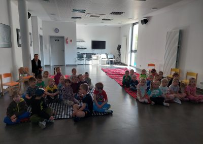 Grupa przedszkolaków siedzi na matach w jasnej sali warsztatowej i słucha prezentacji edukacyjnej. Niektóre dzieci patrzą w stronę prowadzącego, inne już planują mentalną emigrację do świata przekąsek.