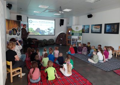 Grupa przedszkolaków siedzi na matach w jasnej sali warsztatowej i słucha prezentacji edukacyjnej. Niektóre dzieci patrzą w stronę prowadzącego, inne już planują mentalną emigrację do świata przekąsek.