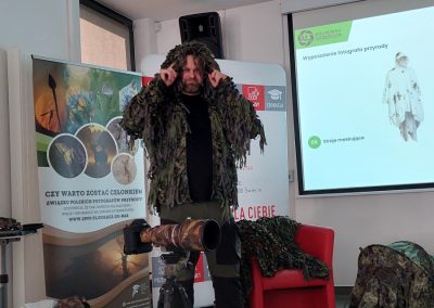 Fotograf przyrody prezentuje strój maskujący przypominający ghillie suit. Stoi obok aparatu z teleobiektywem i pokazuje elementy kamuflażu. Wygląda jak skrzyżowanie leśnego ducha z bardzo zmęczonym krzakiem.