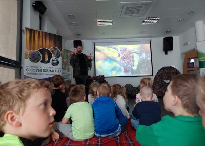Szerokie ujęcie sali podczas warsztatów przyrodniczych. Dzieci siedzą na kocach i oglądają prezentację prowadzoną przez fotografa stojącego przy ekranie.