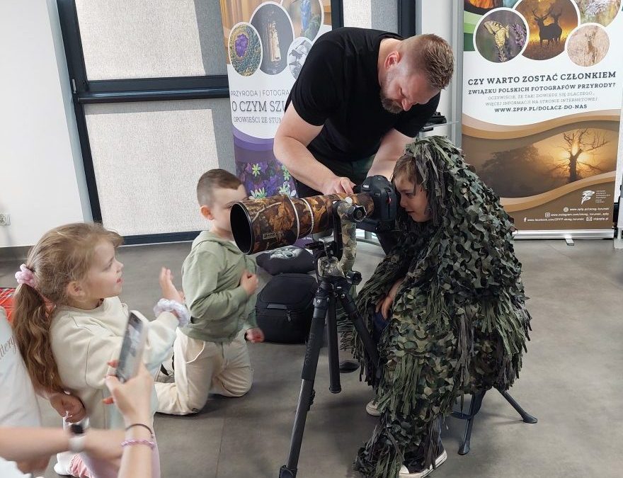 Fotograf pomaga dziecku obsługiwać aparat fotograficzny z teleobiektywem podczas praktycznych zajęć. Inne dzieci obserwują pokaz z bliska.