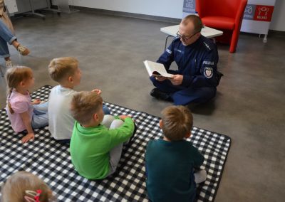 Na zdjęciu widać policjanta siedzącego na podłodze w bibliotece i czytającego książkę grupie małych dzieci siedzących na czarno-białym kocu. Spotkanie odbywa się w jasnej, przestronnej sali bibliotecznej.