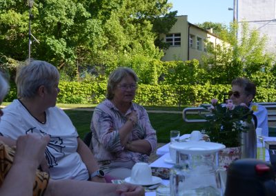 Kobiety siedzą przy stole i rozmawiają podczas spotkania czytelniczego na świeżym powietrzu. Jedna z uczestniczek gestykuluje podczas wypowiedzi, a na stole stoją filiżanki, dzbanek z wodą i kwiaty. W tle widać dużo zieleni i budynki biblioteki.