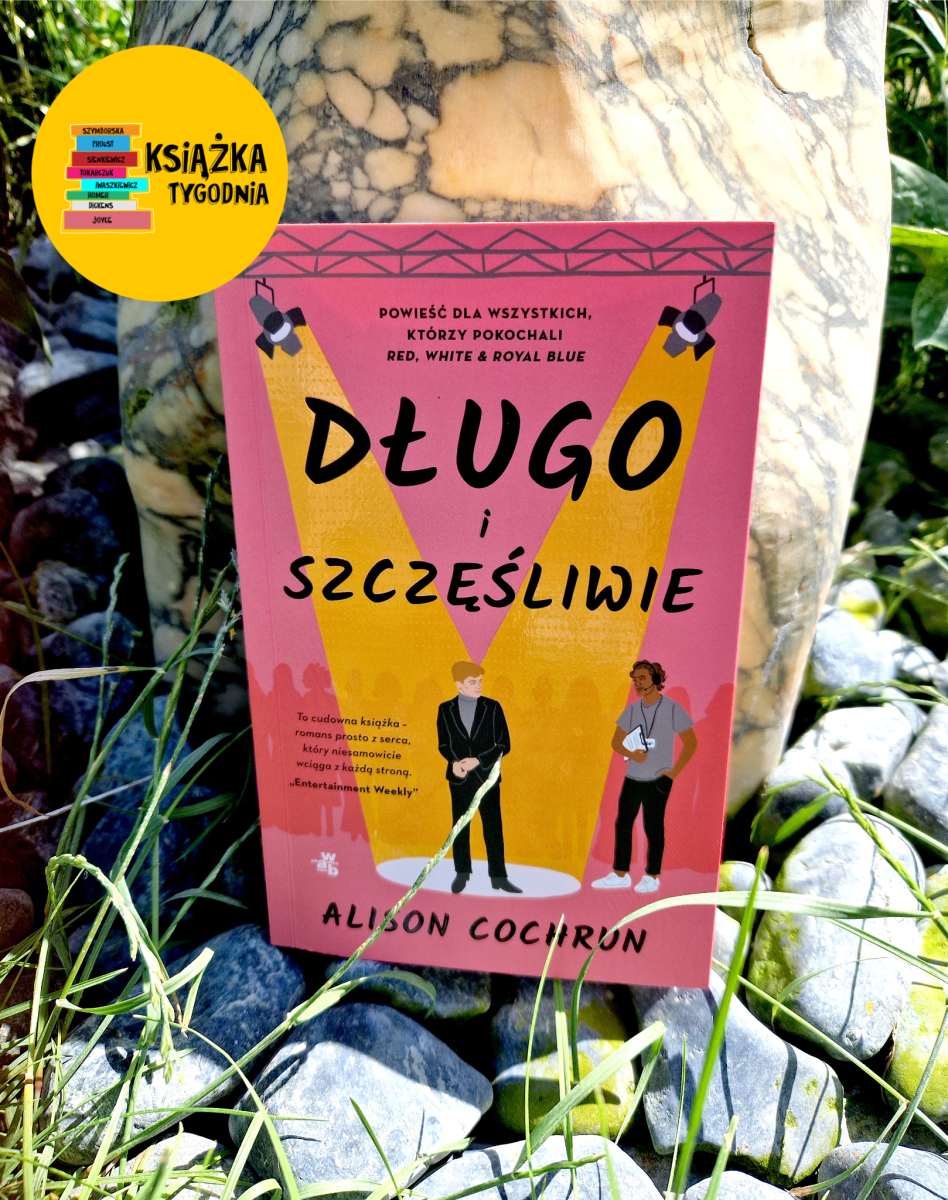 Okładka książki „Długo i szczęśliwie" autorstwa Alison Cochrun, sfotografowana na tle kamiennego głazu i trawy. Okładka ma różowe tło z żółtymi reflektorami scenicznymi skierowanymi na dwie postaci: mężczyznę w czarnym garniturze oraz osobę w szarej koszulce trzymającą notatnik. U góry okładki widnieje napis: „Powieść dla wszystkich, którzy pokochali Red, White & Royal Blue". W lewym górnym rogu zdjęcia znajduje się żółta okrągła naklejka z napisem „Książka Tygodnia" i kolorowymi grzbietami książek z nazwiskami autorów (m.in. Szymborska, Proust, Tokarczuk, Dickens, Joyce). Na dole okładki cytat z „Entertainment Weekly": „To cudowna książka – romans prosto z serca, który niesamowicie wciąga z każdą stroną."