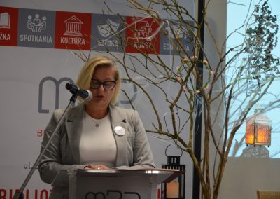 Kobieta w okularach czyta dokument przy mikrofonie na tle gałęzi ozdobionych wiankiem.